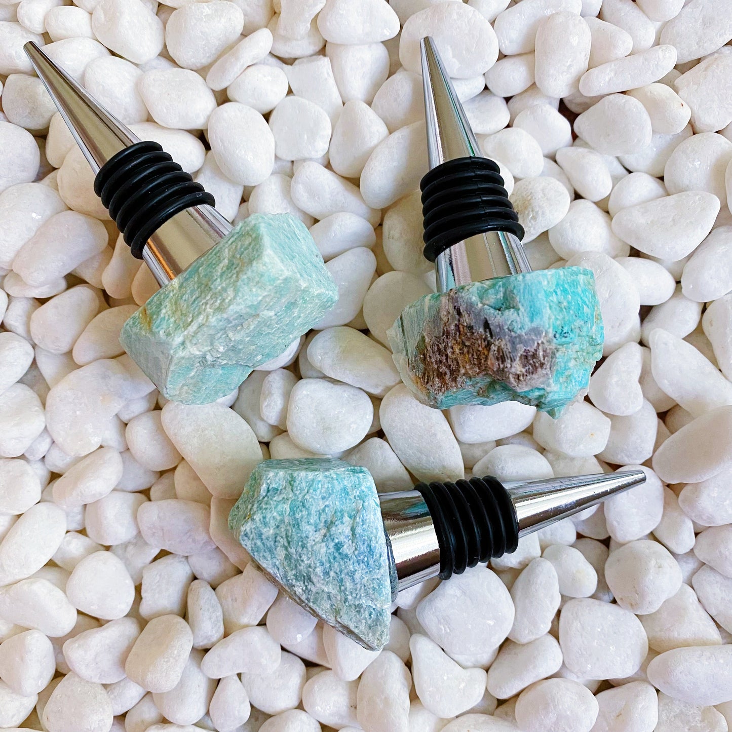 Three wine bottle stoppers with gemstone tops on a bed of white pebbles.