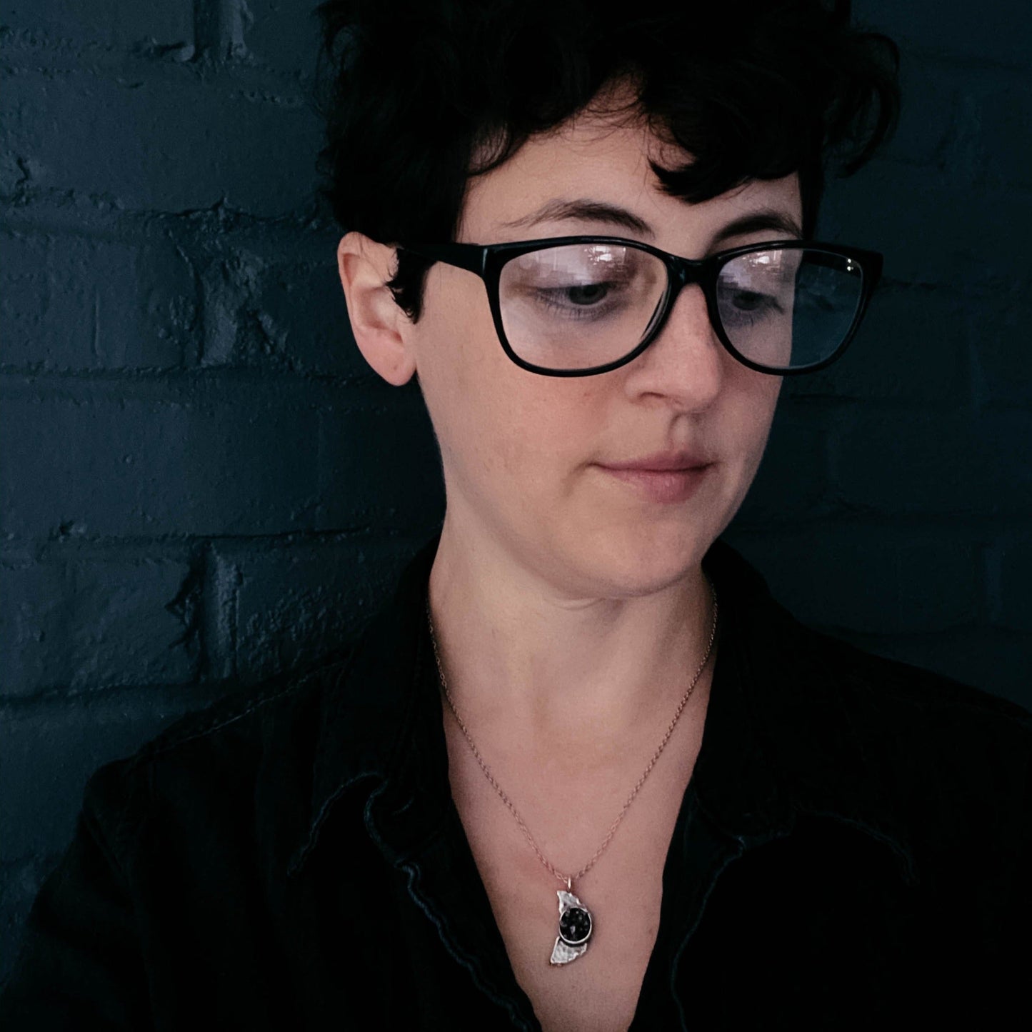 Person wearing a meteorite pendant, glasses and a black shirt against a dark brick wall
