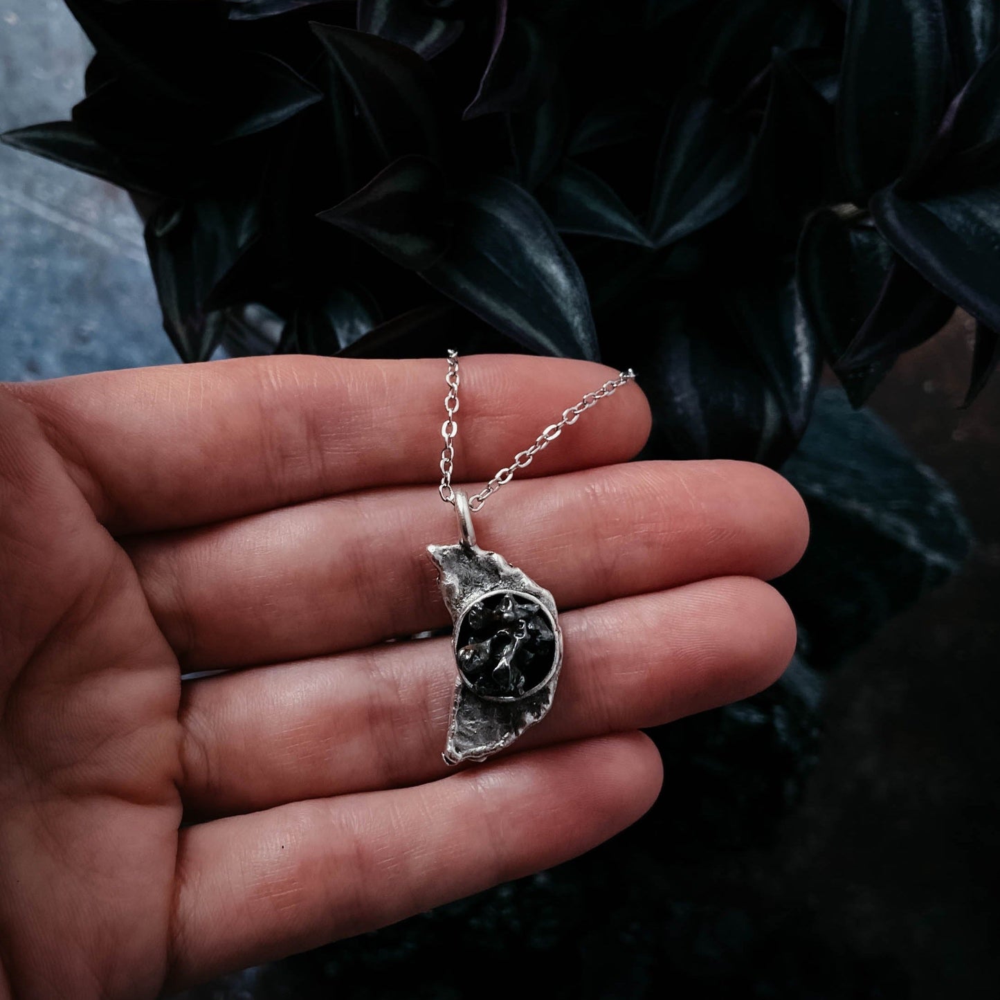 Silver necklace with a meteorite pendant held in a hand against a dark background