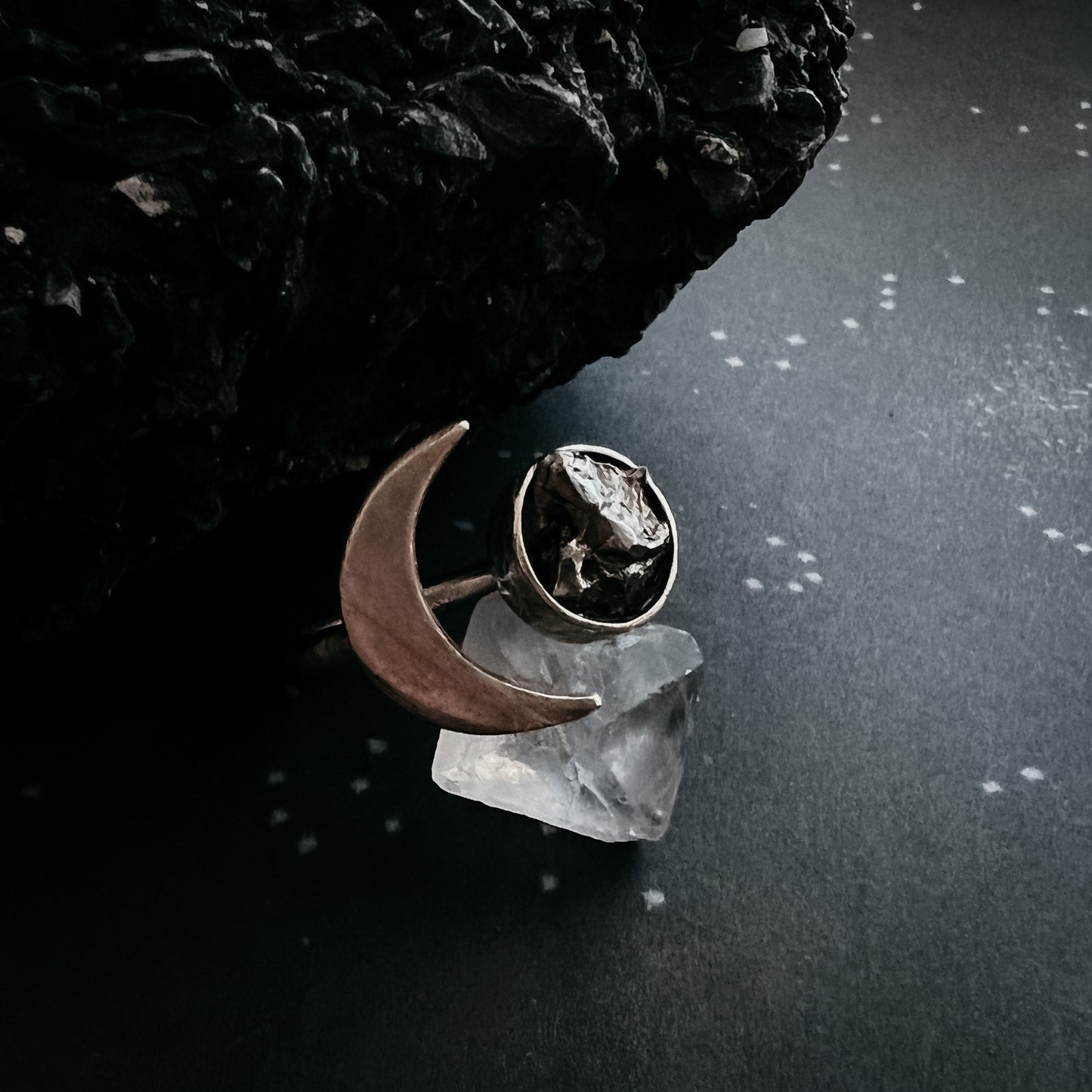Silver ring with meteorite and moon design on a dark surface with a crystal and rock.