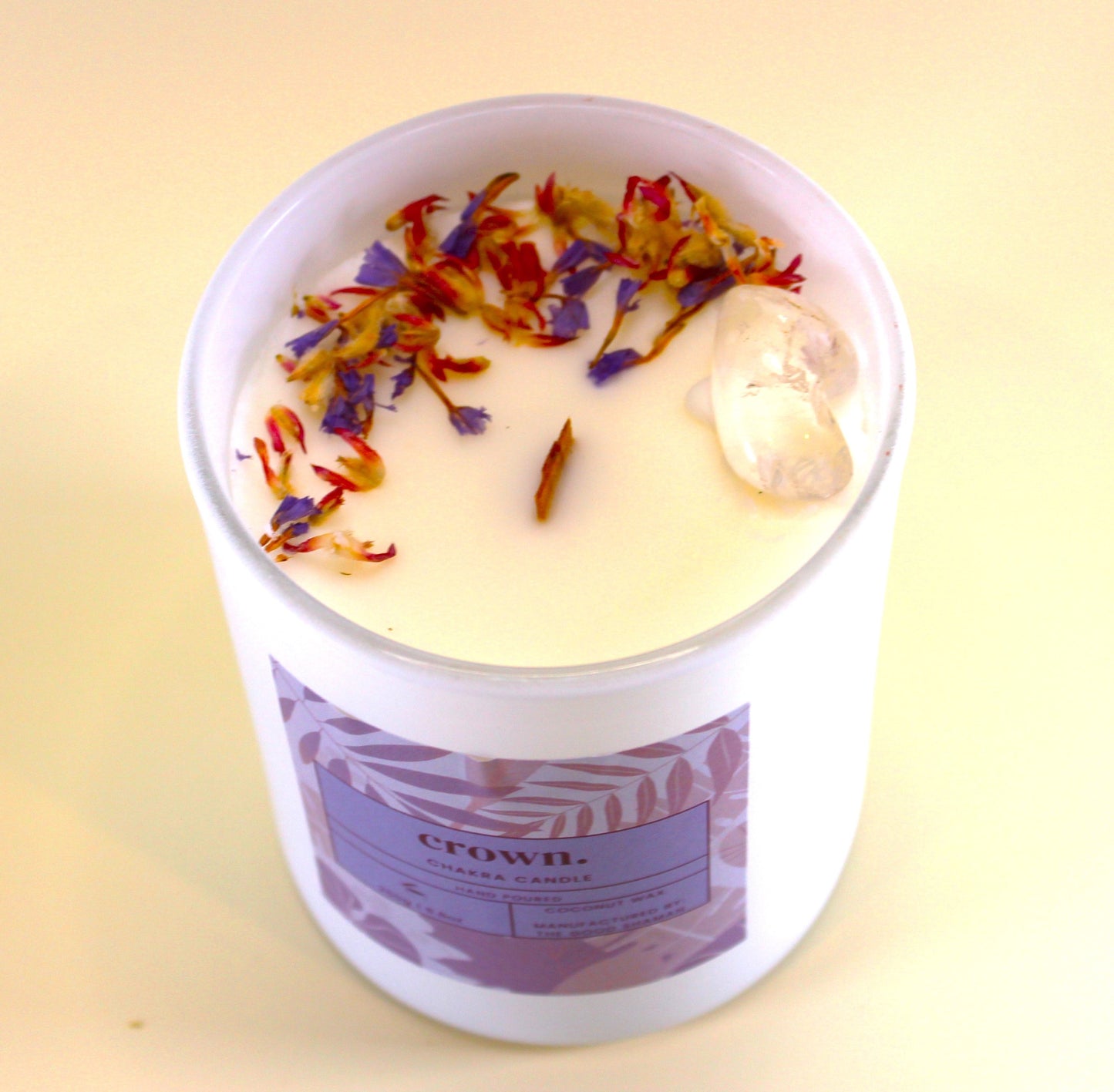 Crown chakra candle in a white container with colorful floral bits on a light yellow background