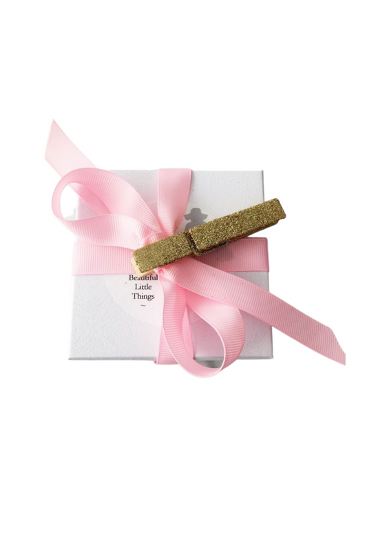 Glittery hair clip on a pink ribbon with a white box labeled 'Beautiful Little Things' gift box on a white background