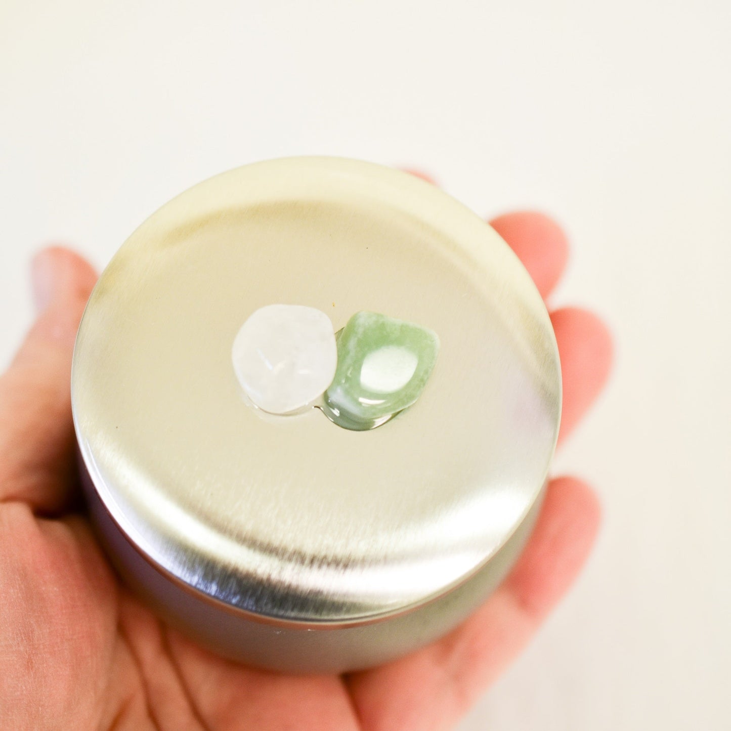 Hand holding a small round candle container with a silver lid and green design on a white background