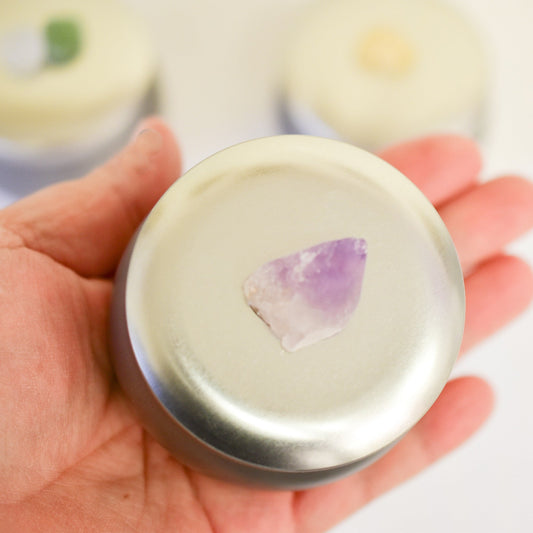 Hand holding a round container with a purple stone on top against a blurred background