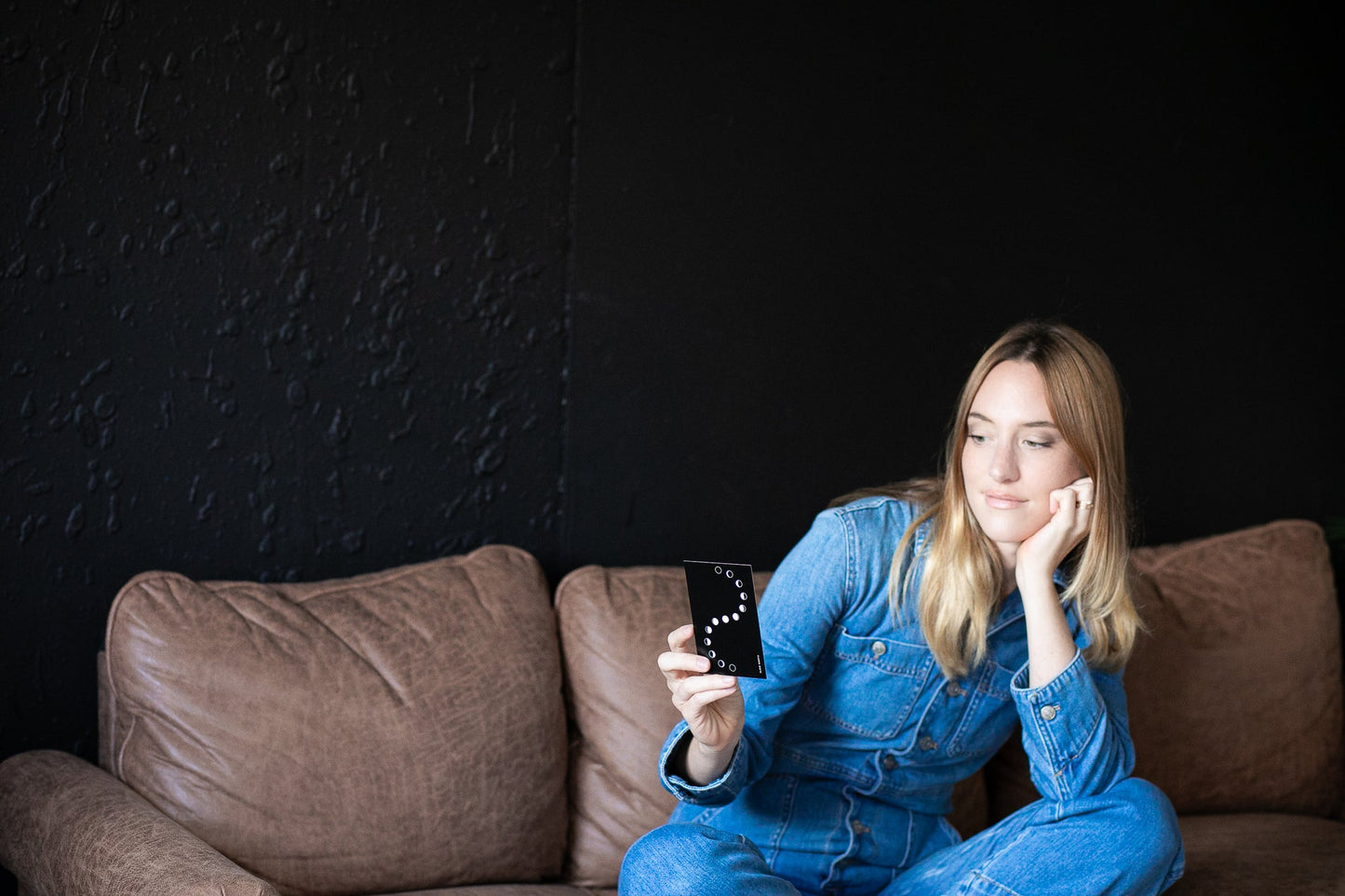 Woman in blue denim outfit sitting on a brown couch holding a smartphone with a dark background