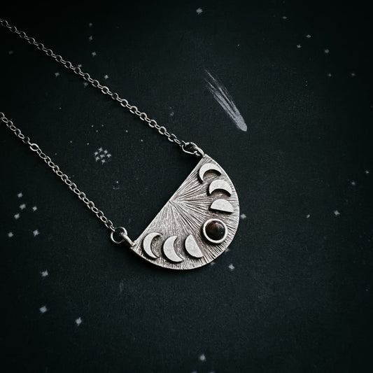 Silver necklace with a half-circle pendant featuring moon phases on a dark background.