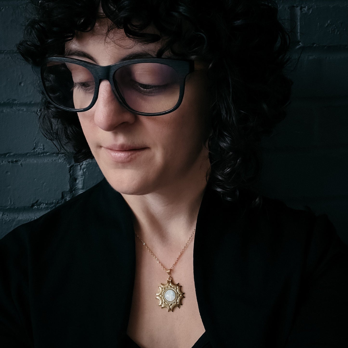 Woman wearing glasses and a black shirt with a gold Moonstone Pendant necklace against a dark background