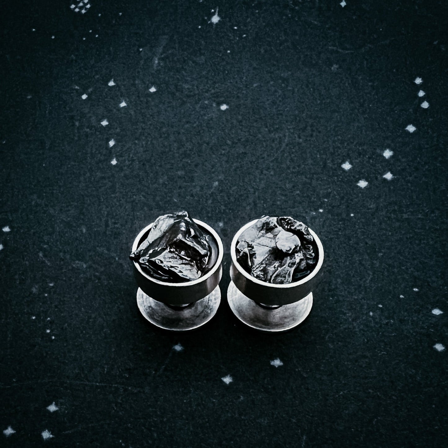 Two meteorite cufflinks on a textured black surface with star-like white dots.
