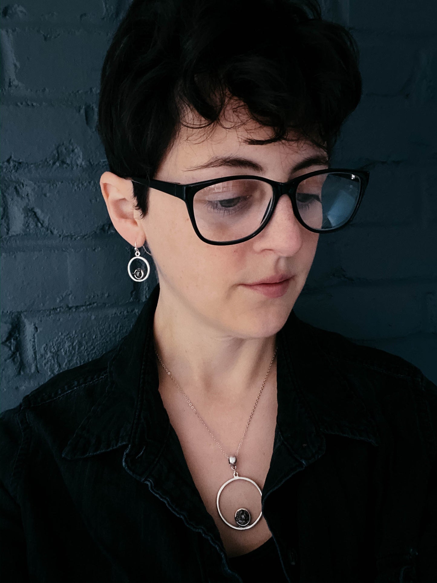 Woman wearing black glasses and a black shirt against a dark brick wall.