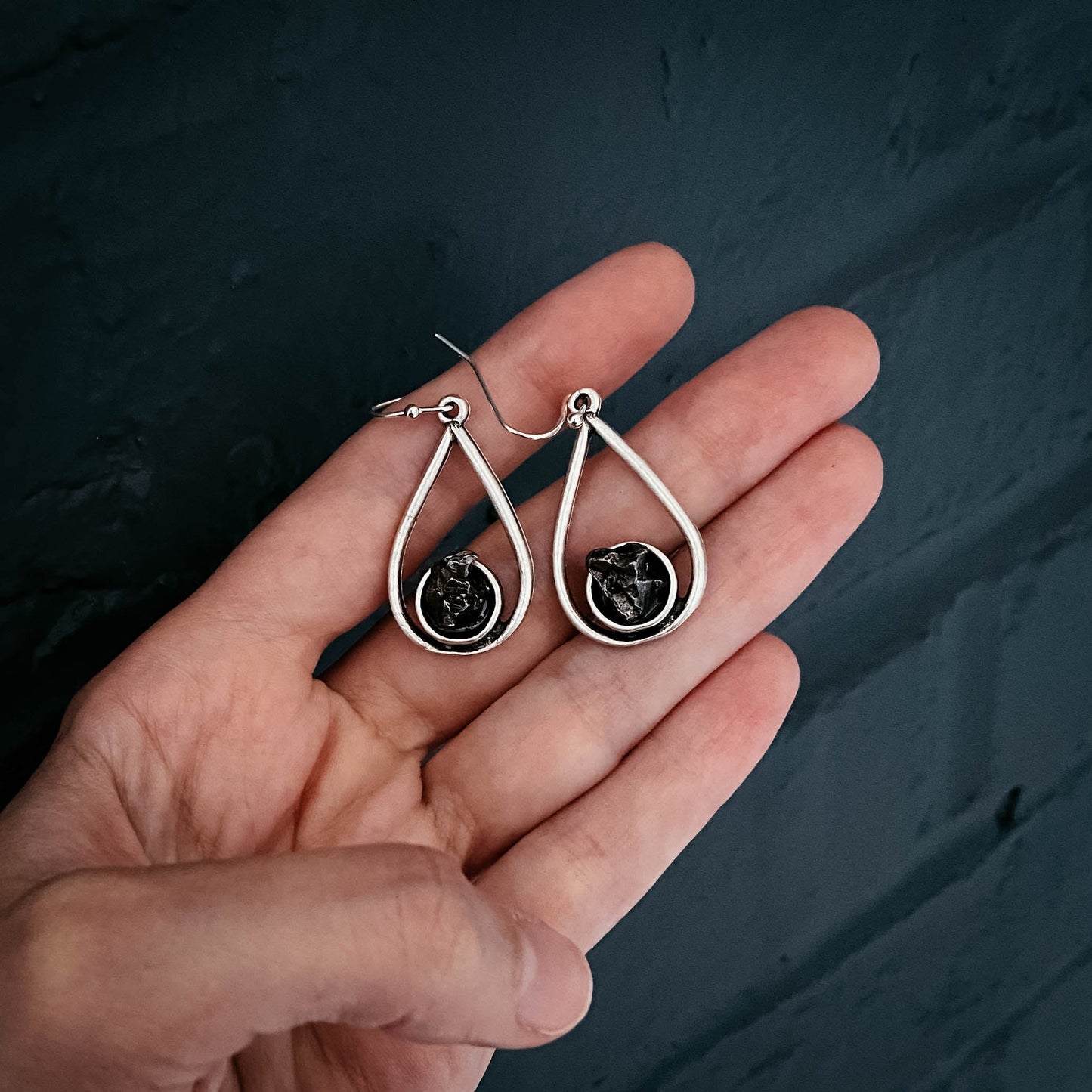 Silver teardrop earrings with meteorite held in a hand against a dark background
