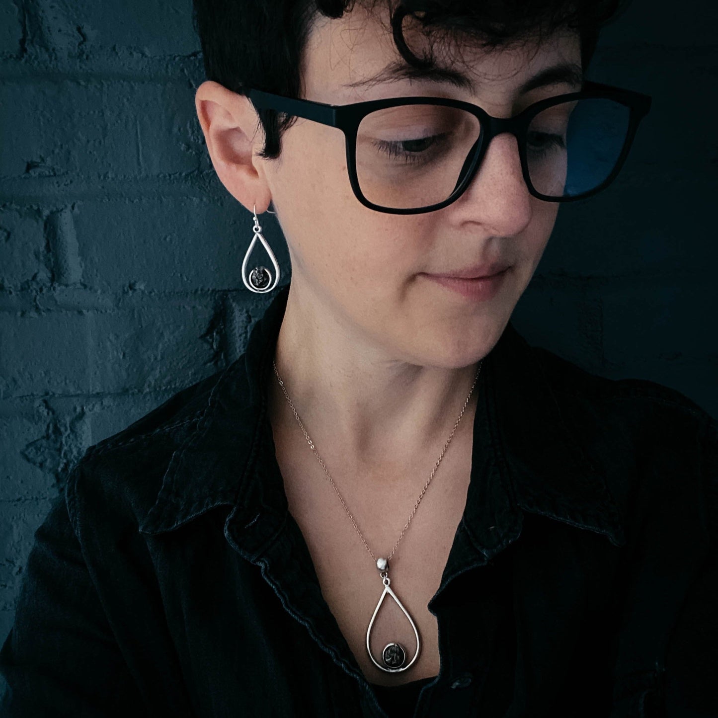 Woman wearing black glasses and a black shirt with silver earrings and necklace against a dark brick wall.