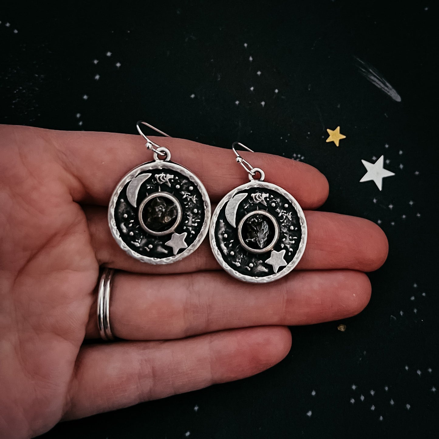 Silver earrings with celestial design held in a hand against a starry background