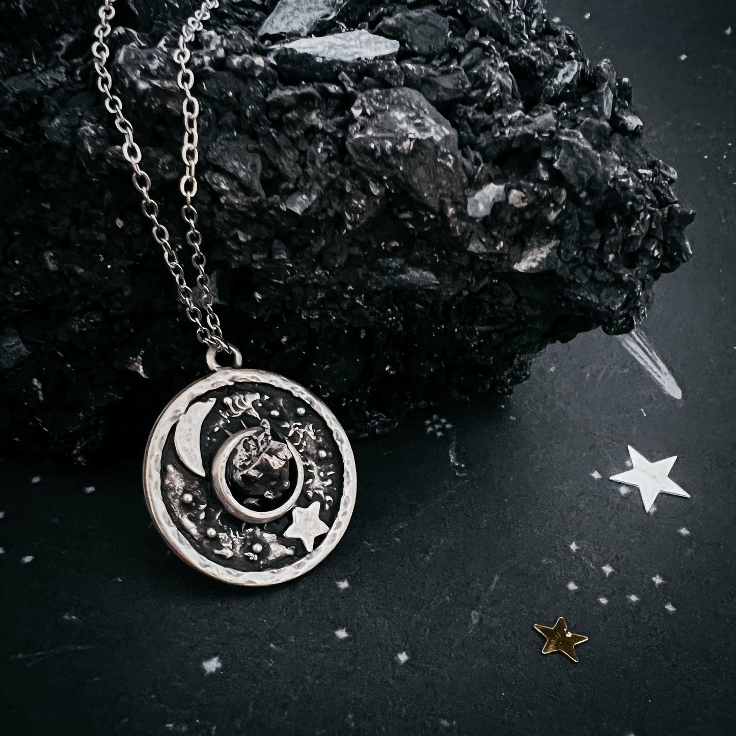 Silver necklace with a moon and star pendant on a dark surface with rocks and stars.