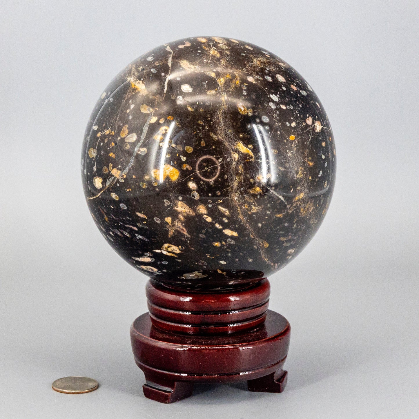 Decorative ocean jasper stone sphere on a wooden stand with a coin for scale on a light gray background