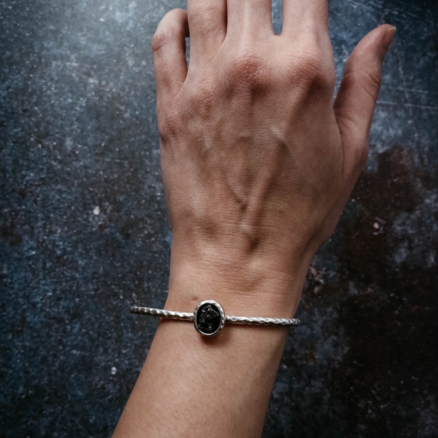 Hand wearing a silver bracelet with a meteorite on a dark textured background