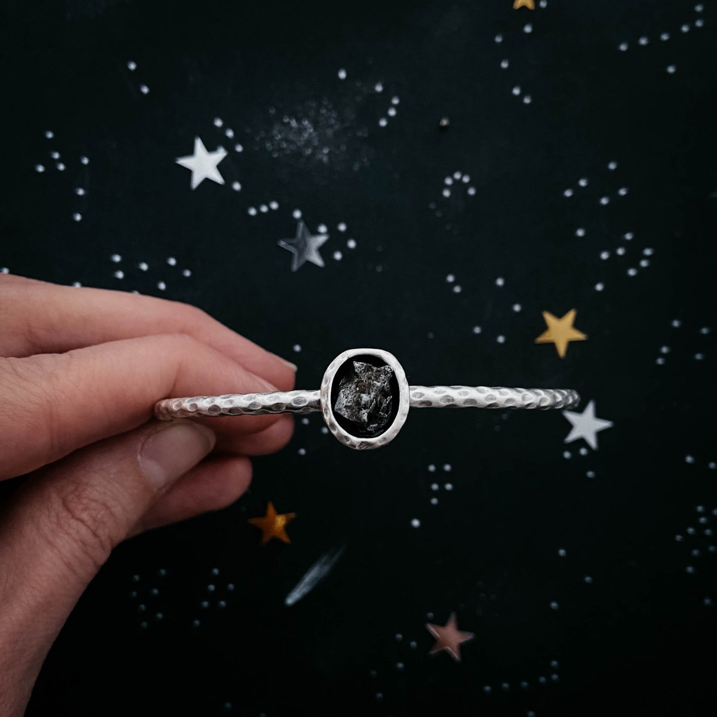 Silver bracelet with meteorite held by a hand against a starry night sky background