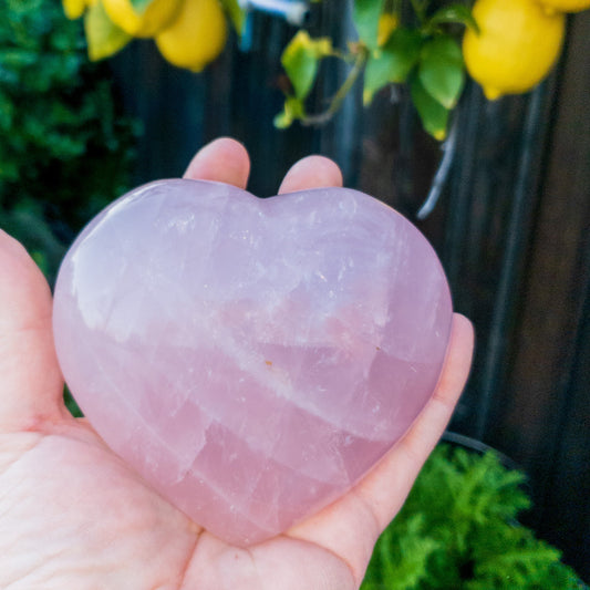 Heart-shaped pink crystal held in a hand with a blurred background of greenery and yellow flowers.