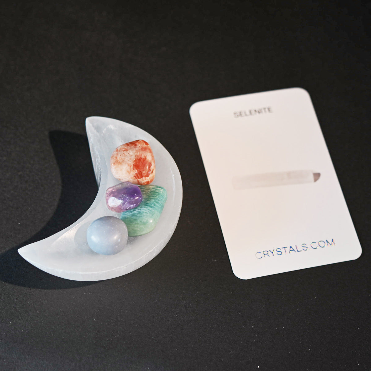 Selenite crescent moon-shaped holder next to a 'Crystals.com' card on a black background.