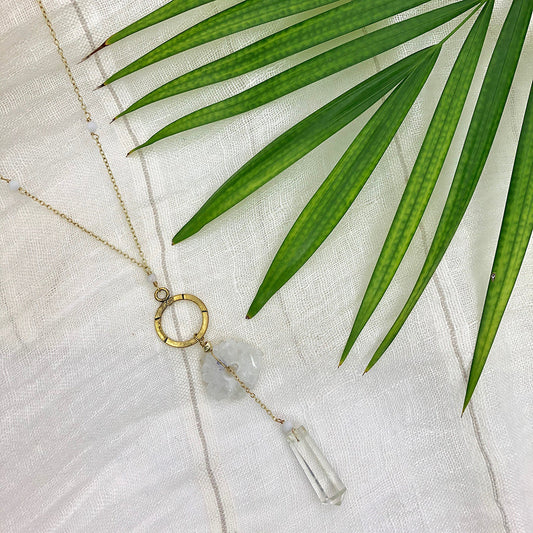 Necklace with a gold ring and clear pendant on a white fabric background with green leaves.