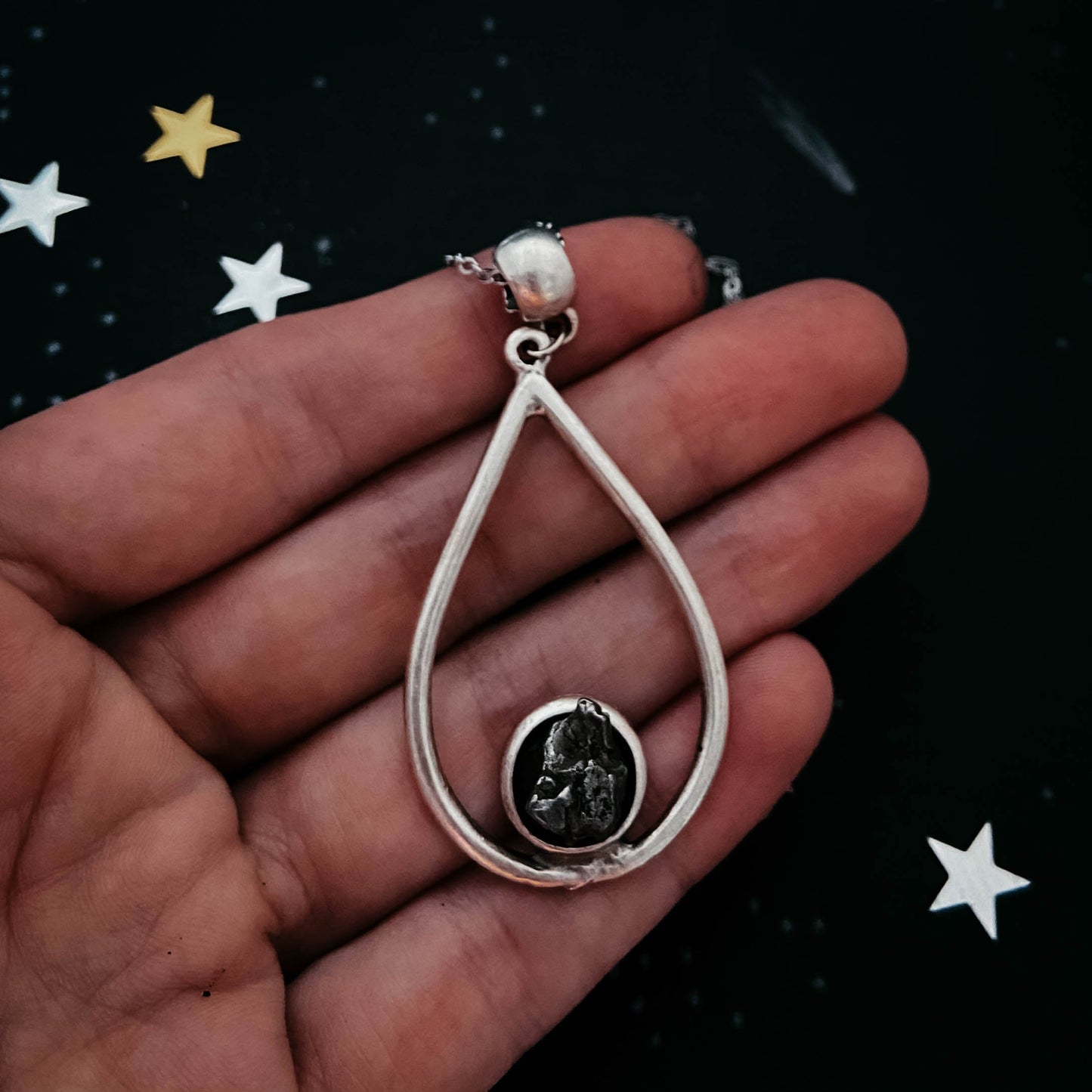 Teardrop-shaped pendant with meteorite held in a hand against a dark background with stars.