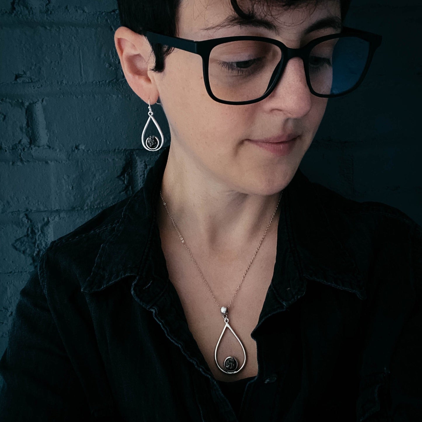 Woman wearing black glasses and a black shirt with a dark background and pendant