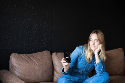 Woman in blue denim outfit sitting on a brown couch holding a smartphone with a dark background