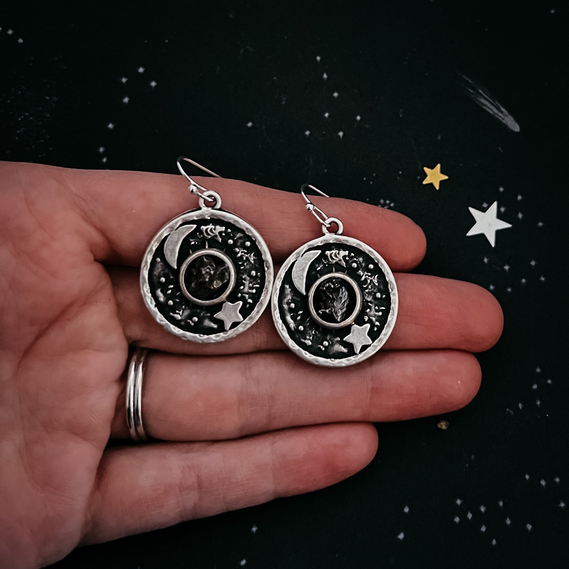 Silver earrings with celestial design held in a hand against a starry background