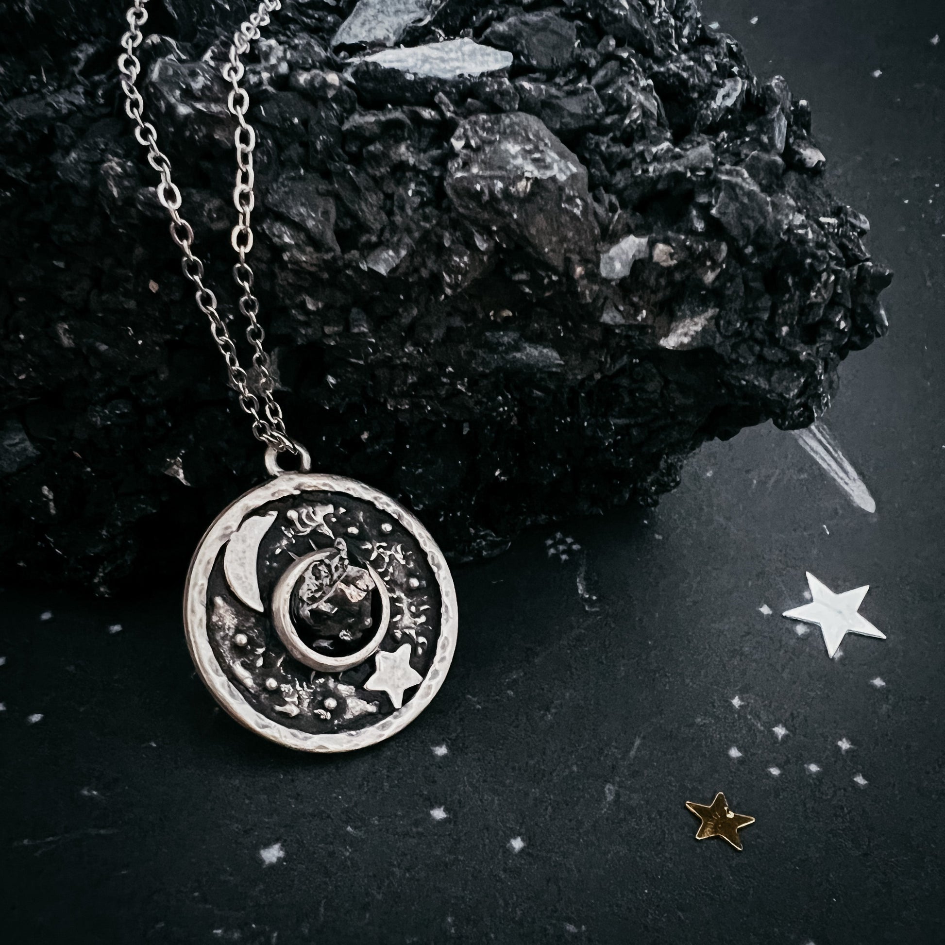 Silver necklace with a moon and star pendant on a dark surface with rocks and stars.