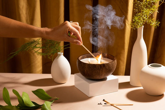 Person lighting a soy wax coconut shell candle with a match, surrounded by decorative items on a table.