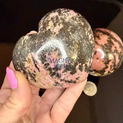 Heart-shaped rhodonite stone held by a hand with pink nail polish.
